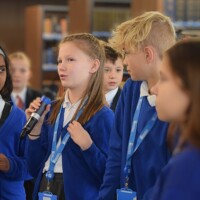 A group of children in blue uniforms, wearing lanyards, are engaged in conversation. One child is speaking into a microphone while others listen attentively.