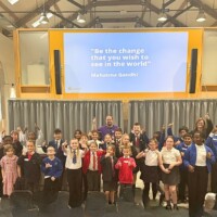 A group of adults and primary school age children pose in front of a screen displaying a quote by Mahatma Gandhi in a hall. The adults and many of the children are holding their thumbs up at the camera and are smiling.
