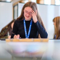 A girl with long hair and glasses and wearing a blue lanyard, writes with a pen at a table. Other people and blurred background are visible.