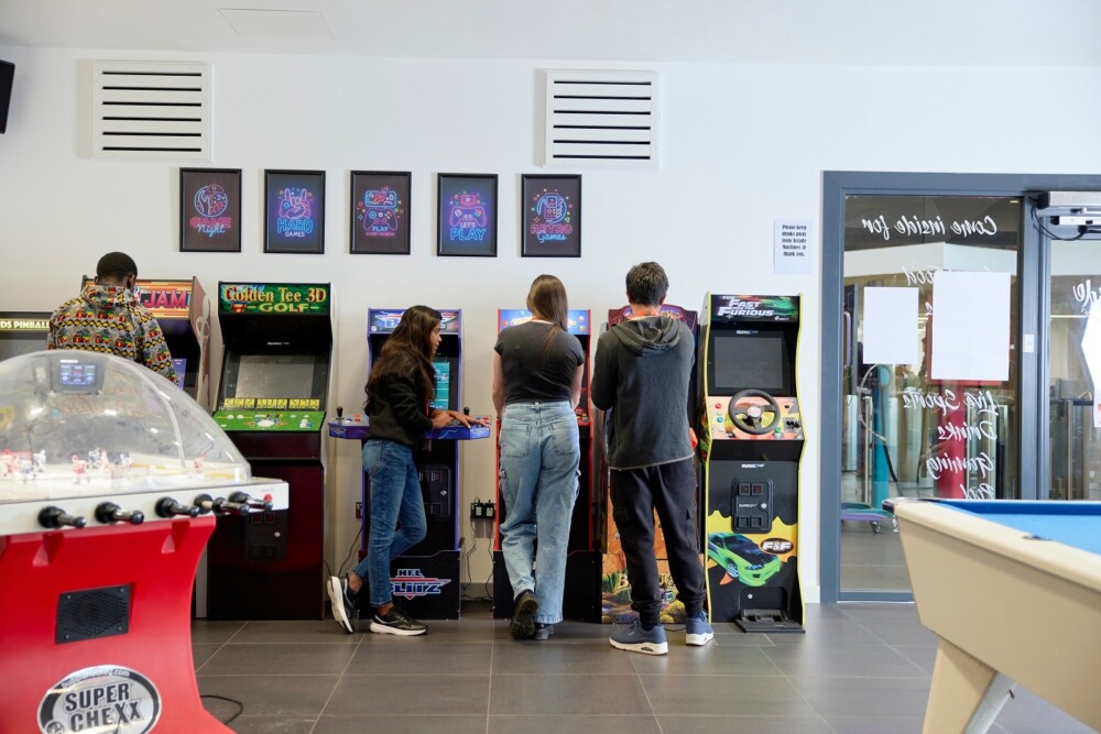 Students play arcade games in a game room, with a variety of machines lined up along the wall. In the foreground, there is a snooker table.