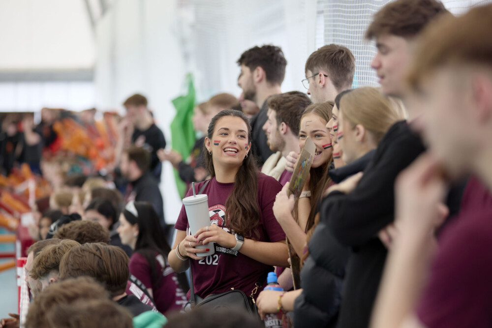 A crowd of students in a stadium, with a woman in a maroon shirt holding a white travel cup and smiling. Others around her are engaged in conversation.