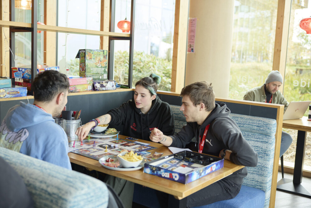 Three students playing a board game in a casual cafe setting with snacks and drinks on the table. A person in the background is using a laptop.