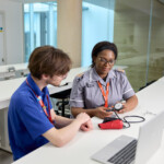 Two Learning Disability Students in a modern office setting, discussing a blood pressure monitor at a table with a laptop.