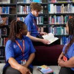 A group of three Learning Disability students in blue uniforms are sitting in a library, with one person standing and arranging books. Two seated individuals listen to another person reading from an open book.