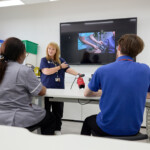 A Learning Disability academic is standing in front of a screen with an image of a nurse conducting a blood pressure test, while also holding out their own arm to demonstrate to two students sitting at white benches.