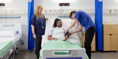 A patient lies in a hospital bed while a Learning Disability Nursing student and an academic monitor her.