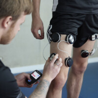 Person adjusts electrical stimulation devices on another person's leg using a handheld controller in a gym setting.