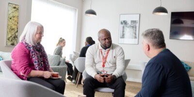 Three people seated and conversing in a lounge area with overhead lighting and framed artwork. They hold cups, and more people are visible in the background.
