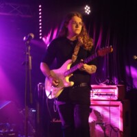 A guitarist with long hair plays an electric guitar on stage under purple lighting, next to a microphone and amplifier.
