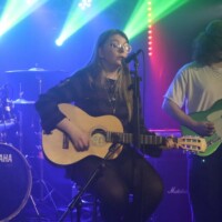A band performs on stage with colorful lights. A woman plays an acoustic guitar and sings, while two men play the drums and electric guitar.