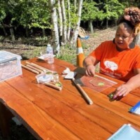 A woman in an orange shirt works on a nature craft project at a wooden table outdoors, with various supplies and trees in the background.