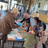 Adults and children gather around a table indoors, engaging in a hands-on activity with dough and cooking materials.