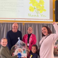 Four adults and two children pose and smile with a gift basket in front of a presentation screen displaying a thank you message and a list of charities.
