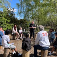 A group of children and adults sit on tree stumps in a circle outdoors, listening to a person standing and speaking on a sunny day.