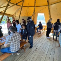 People of various ages interact and participate in activities inside a large teepee with benches, tables, and natural light.
