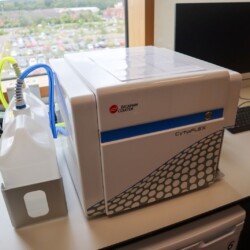 A Beckman Coulter CytoFLEX flow cytometer sits on a lab desk beside a computer monitor, keyboard, and a large plastic container.