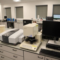 Laboratory workspace with scientific instruments, microscopes, computer monitors, and various lab equipment arranged on countertops and desks.