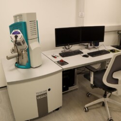 A scanning electron microscope labeled "TESCAN VEGA3" is positioned next to a workstation with dual monitors, keyboard, mouse, and an office chair in a laboratory setting.