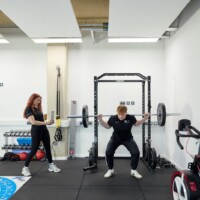A person performs a barbell squat on a squat rack while another person stands nearby observing in a gym setting with weights and exercise equipment.