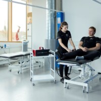 A woman examines a man's leg as he sits on a treatment table in a medical clinic room with additional equipment and beds visible.