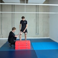 Two men in sportswear prepare for a test involving red foam blocks and sensors attached to one man's legs in a gym.