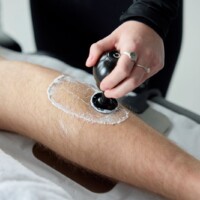 A person applies gel and uses a black handheld device on a person's lower leg as part of therapy treatment plan.