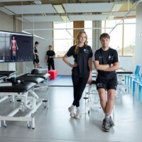 Two students in sportswear stand in a modern clinic with examination tables, blue chairs, and a screen displaying a human anatomy diagram.