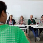 A group of four adults sit at desks facing an instructor in a classroom, with notebooks, a laptop, and water bottles in front of them.