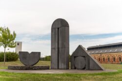 Outdoor sculpture composed of geometric shapes, including a semicircle, a tall rectangle, a triangle, and a circle, set on grass with buildings and a tree in the background.
