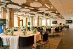 A modern banquet hall set with round tables covered in white cloths, each with folded napkins, glassware, and chairs, prepared for an event or reception.
