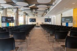 Rows of empty black chairs face a stage with two screens displaying “Work. Play. Stay.” in a modern conference room with ceiling panels and large windows.