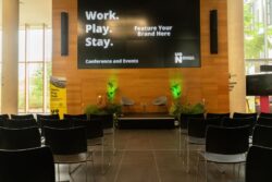 A conference room with black chairs facing a stage, two chairs, plants, and a large screen displaying "Work. Play. Stay. Feature Your Brand Here. Conference and Events.