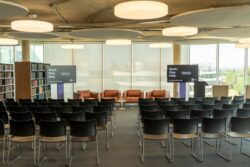 Rows of black chairs face a stage with three brown sofas, a table, and two presentation screens in a modern, well-lit event space with large windows and bookshelves.