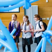 Three schoolgirls in uniform with backpacks look at a display of large blue balloon structures in an indoor setting.
