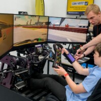 UON 12.06.24 A boy sits in a racing simulator cockpit with three screens, holding a steering wheel, while an adult instructs him.