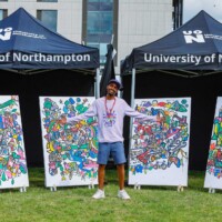 A person stands smiling with arms outstretched in front of four colorful artworks, beneath University of Northampton tents at an outdoor event.