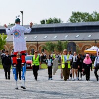 A performer on stilts in a white coat juggles outside as a group of students and staff in high-vis vests walk toward him on a paved area with brick buildings in the background.