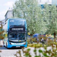 A blue double-decker bus labeled "STEAM Northants 16" is stopped on a tree-lined street with a few people walking nearby.