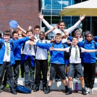 A group of schoolchildren in uniforms stand outdoors with their arms raised, smiling at the camera, with more students and a building in the background.