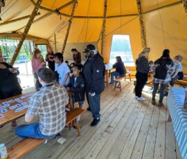 A group of people varying in ages stood inside a tipi within a food education workshop, conversing with one another
