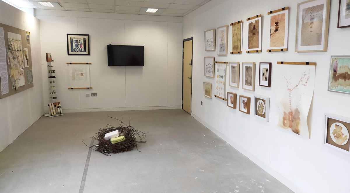View of exhibition with board and display rack with pigments on left, nest with pastel coloured bricks in on floor and 21 works displayed on wall on right