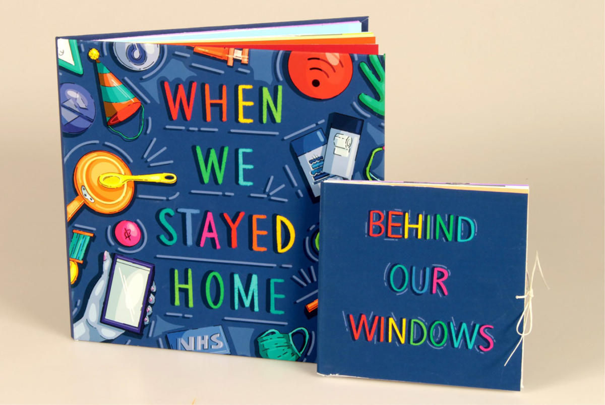A photo of two books. The main book is titled When We Stayed Home, and the other is titled Behind Our Windows Both books are dark blue with multi coloured illustrations and text, situated on a plain beige background.